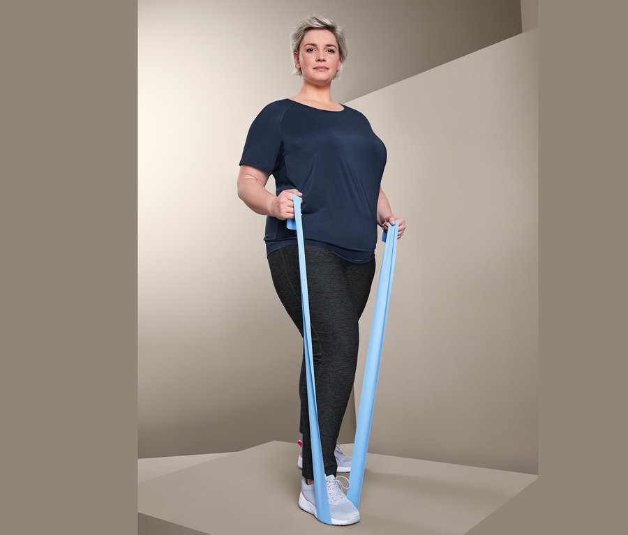 Žena cvičí s modrou posilovací gumou. Má na sobě sportovní triko a sportovní kalhoty.