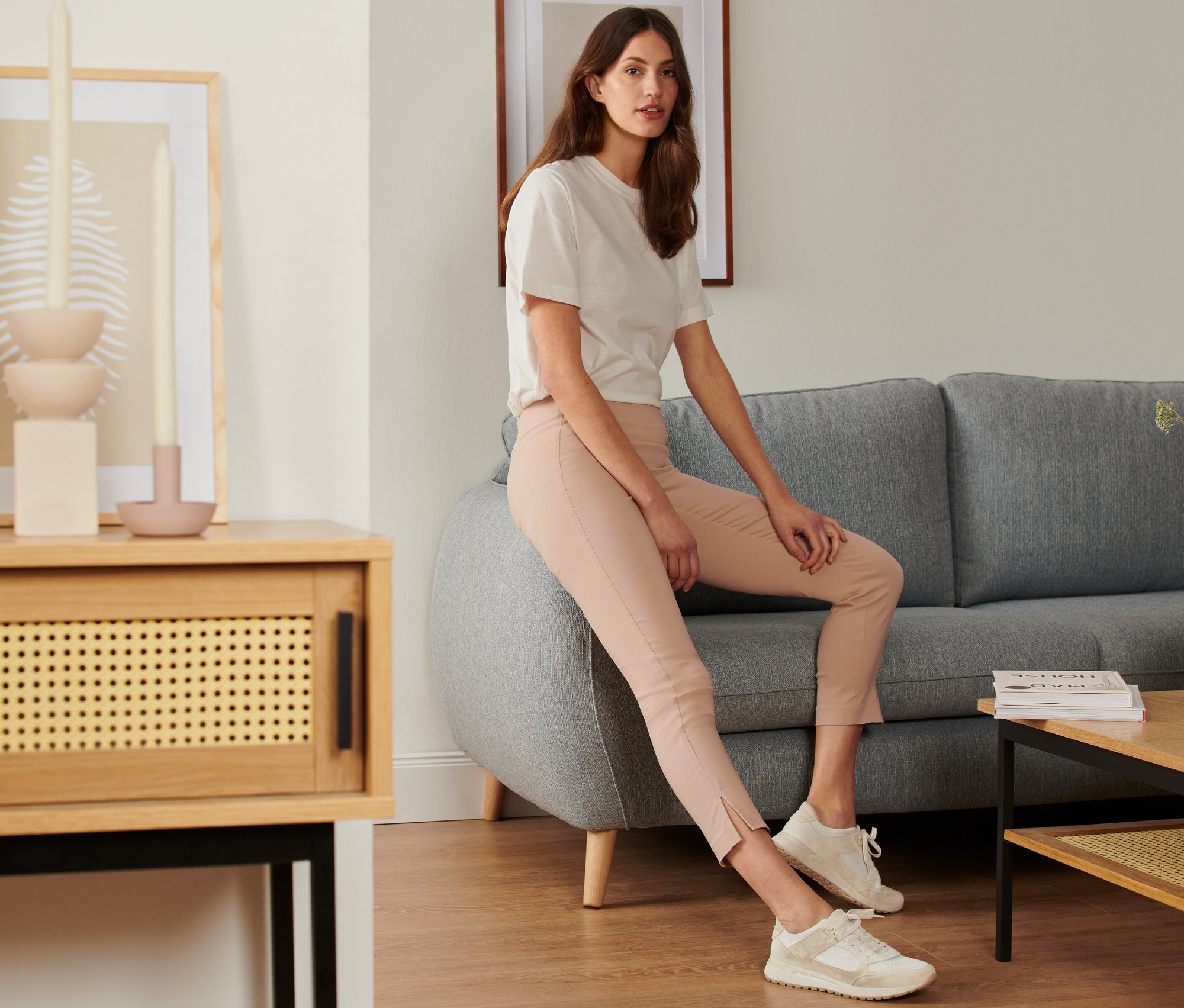 Žena sedí na šedé pohovce a má na sobě béžové strečové kalhoty.