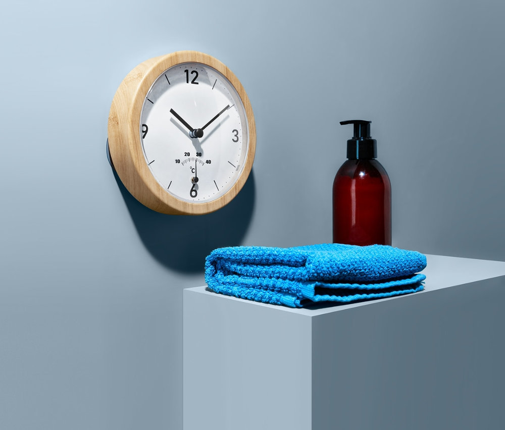 Na zdi visí koupelnové hodiny s teploměrem. Na podstavci leží modrý ručník a hnědá láhev s pumpičkou.