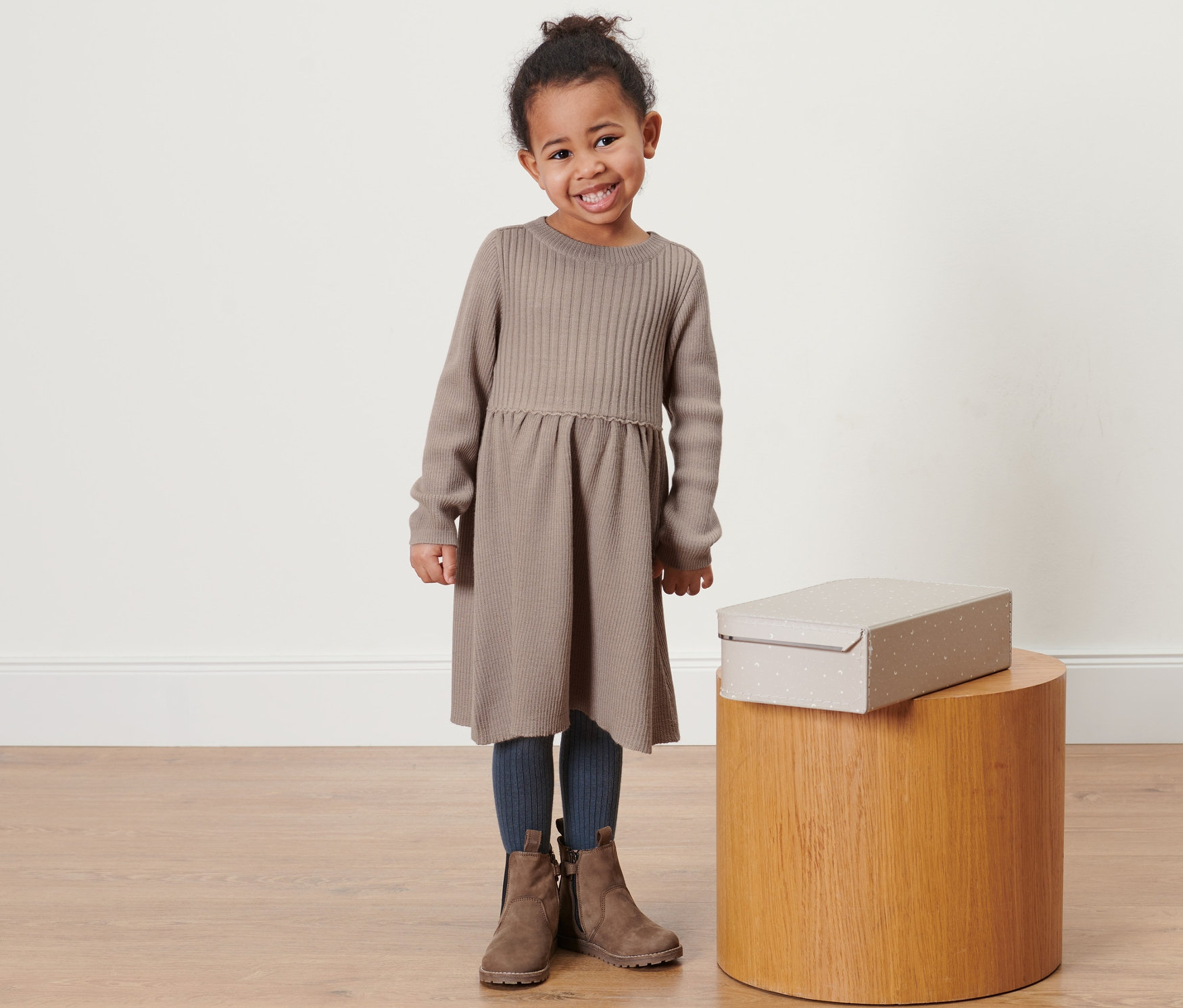 Usmívající se holčička pózuje v pletených šatech merino světle hnědé barvy. Vedle ní stojí na dřevěném stolku krabice.