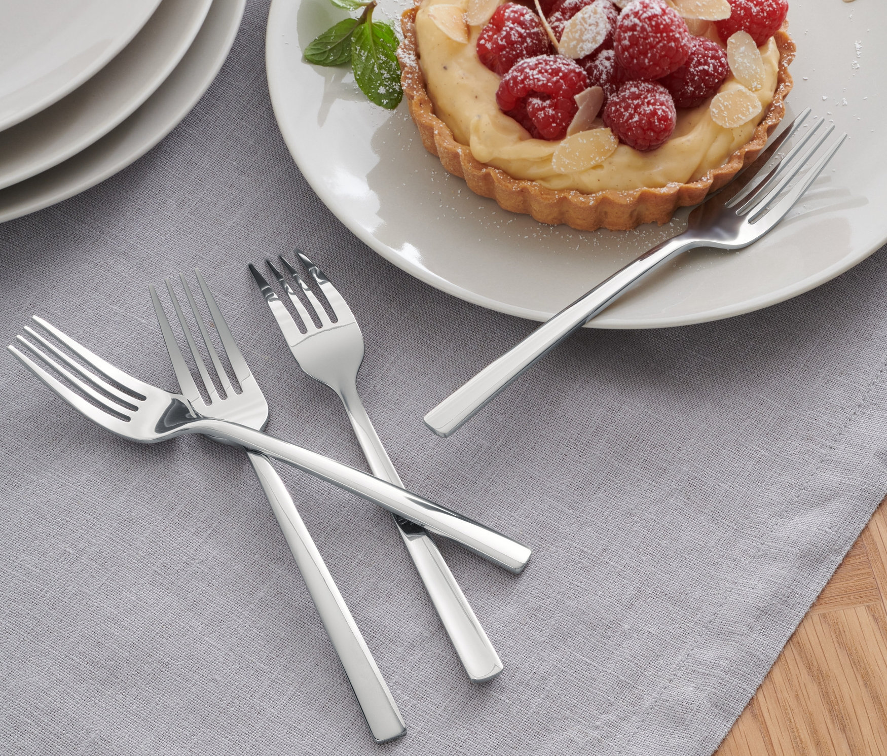 Tři ze 4 vidliček na koláče leží na šedém ubruse vedle talíře s malinovým koláčem.
