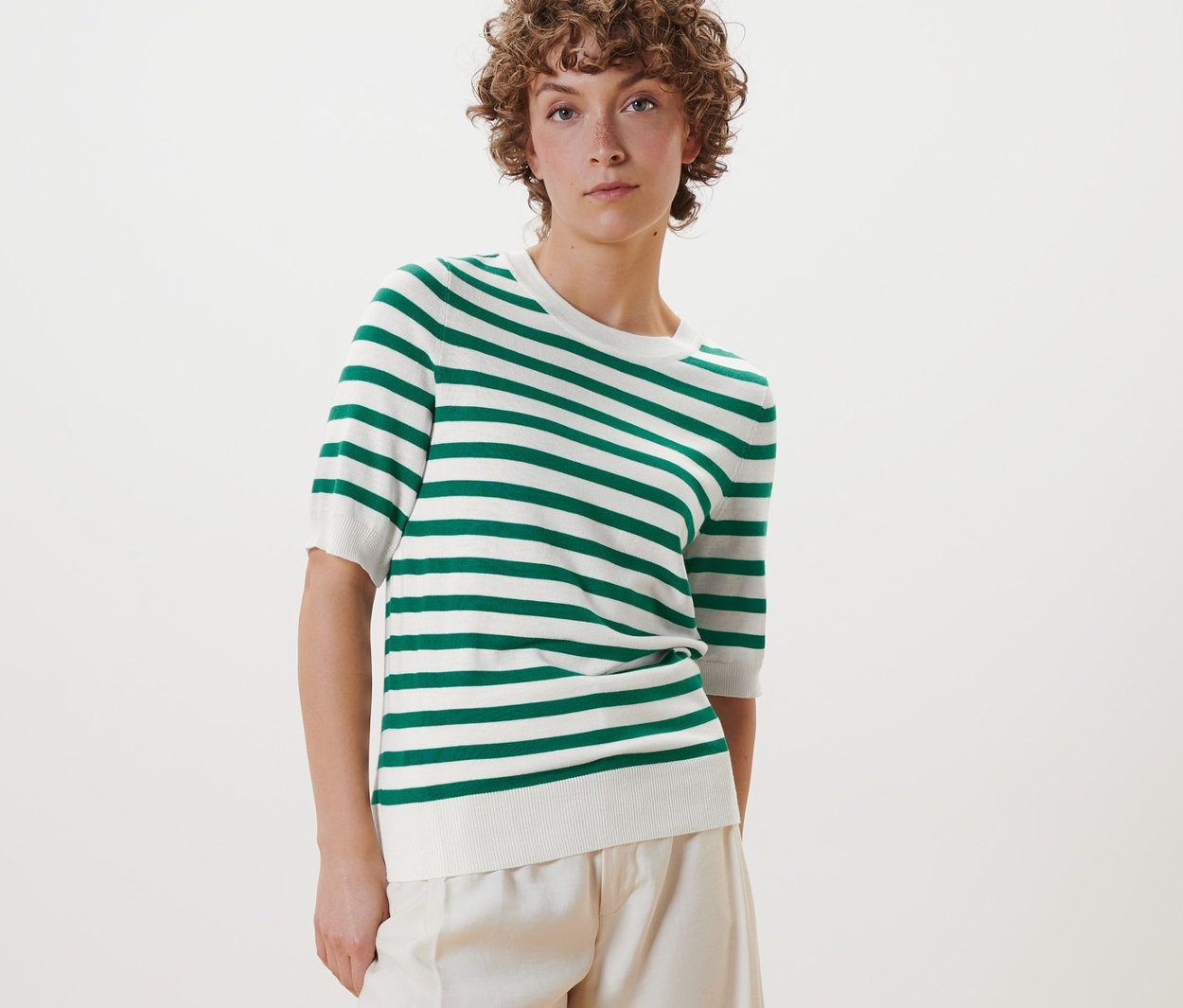 Žena pózuje v pruhovaném svetru z jemného úpletu, bílých kalhotách a bílých botách.