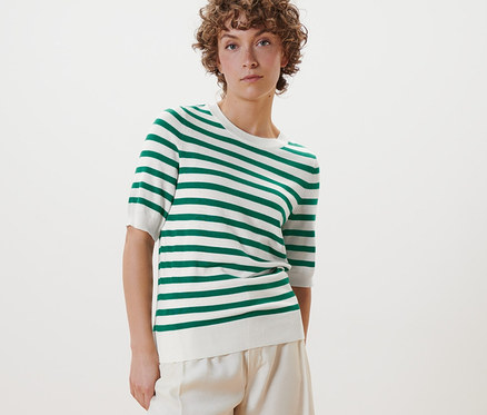 Žena pózuje v pruhovaném svetru z jemného úpletu, bílých kalhotách a bílých botách.