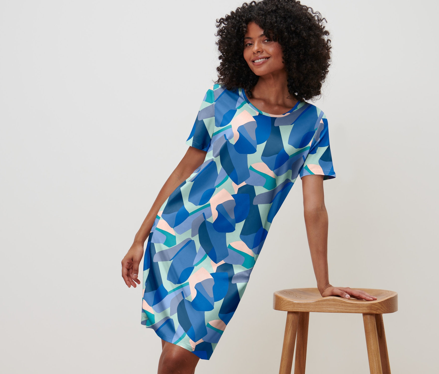 Žena pózuje v modré noční košili s potiskem a opírá se o dřevěnou stoličku.