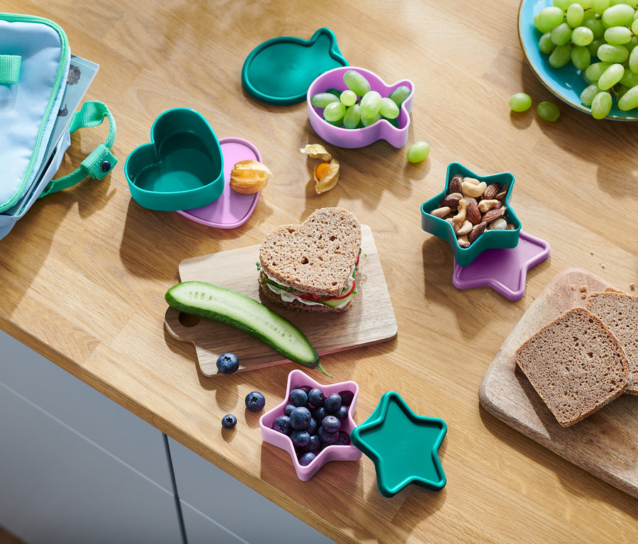 Na dřevěném stole je svačina. Srdcový sendvič, okurka, borůvky v nádobě ve tvaru hvězdy, ořechy, hrozny v nádobě ve tvaru ryby a chléb.