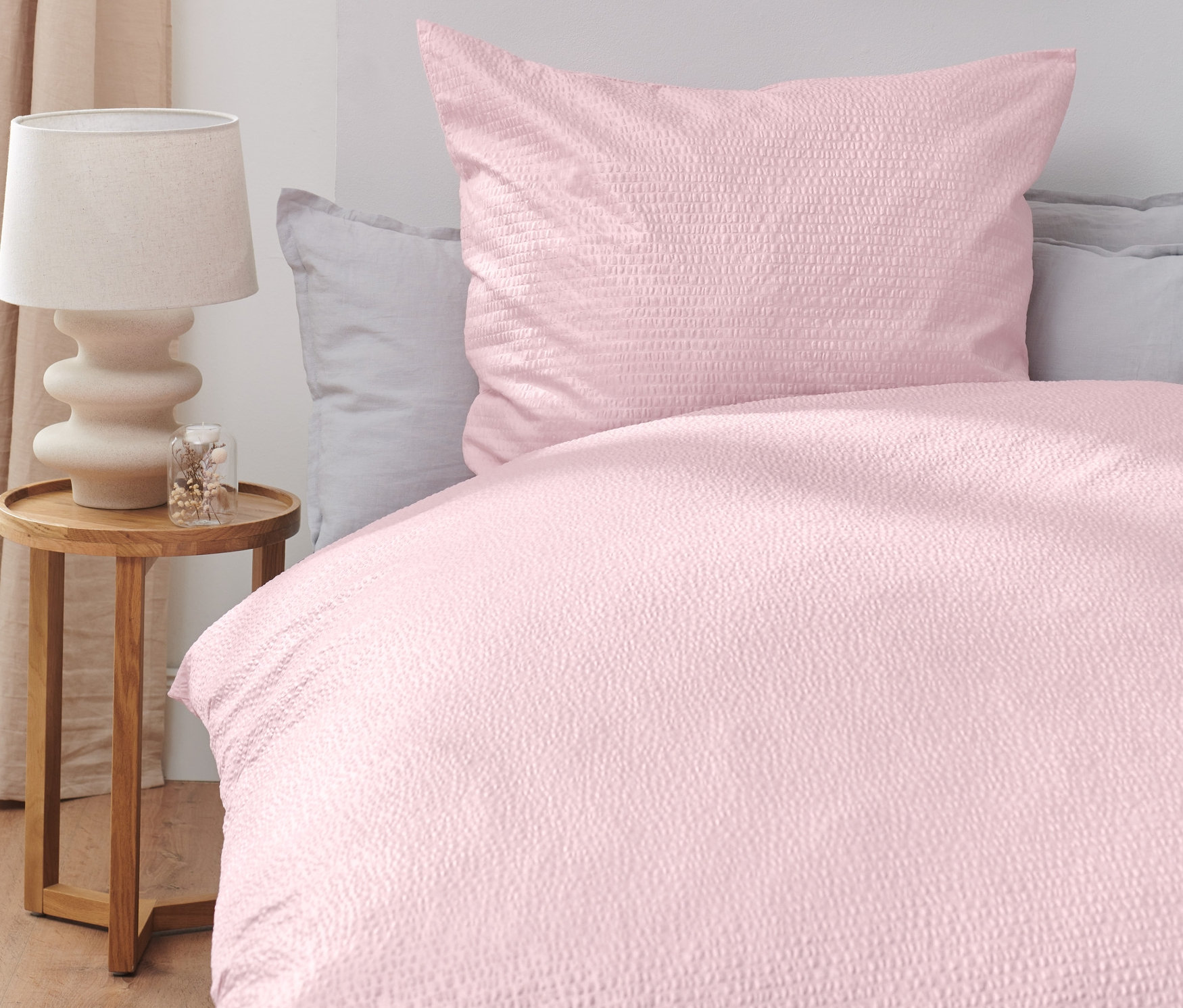 Růžové krepové povlečení na posteli, vedle dřevěný stolek s lampou.