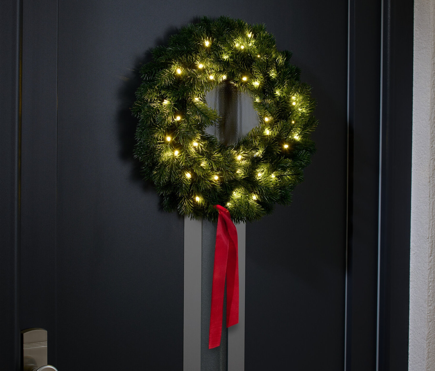 Na černých dveřích visí jedlový věnec s LED světly a červenou stuhou.