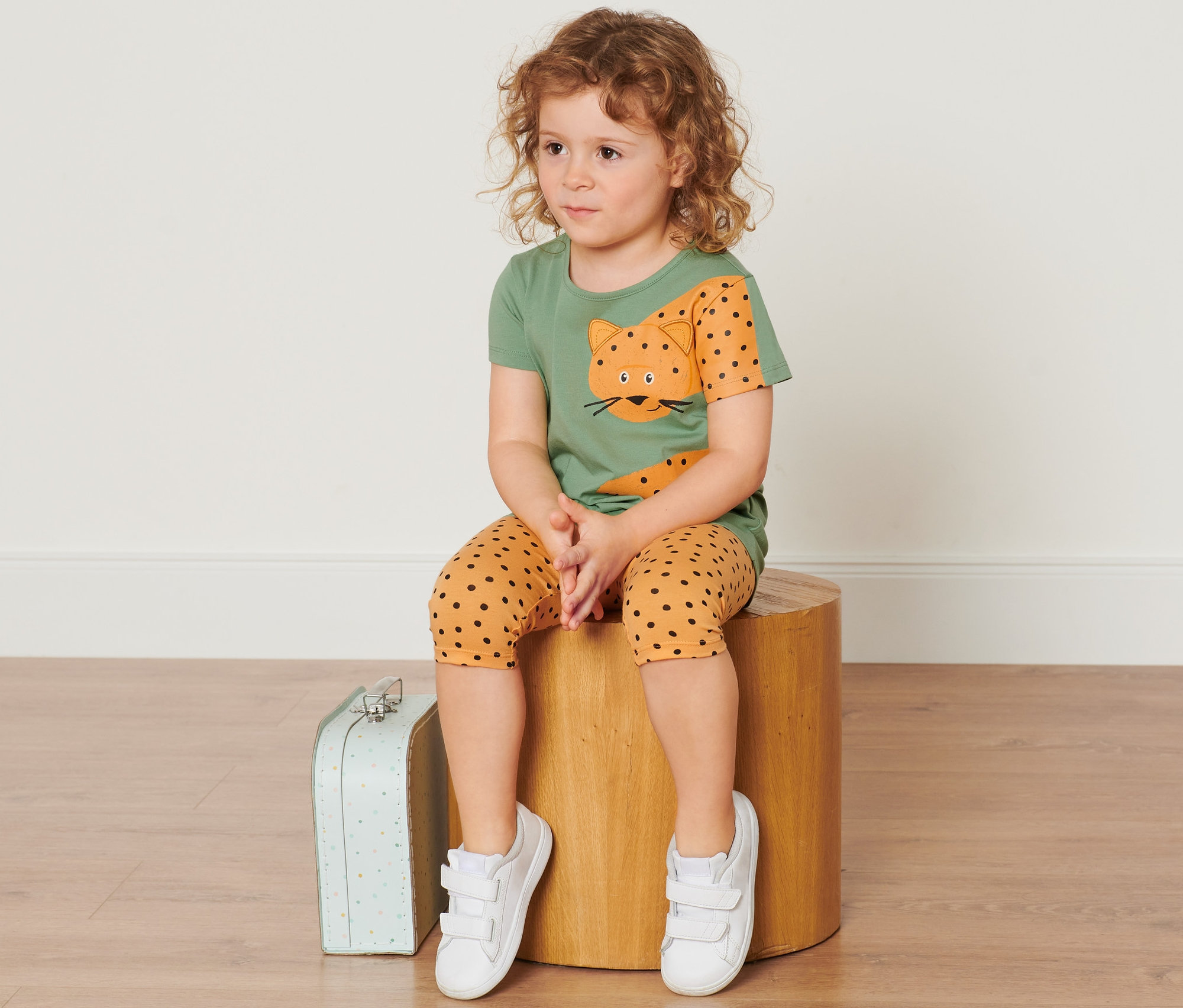 Dítě sedí na dřevěném špalku. Má na sobě zelené tričko a žluté legíny s černými puntíky. Vedle něj leží malý kufr.