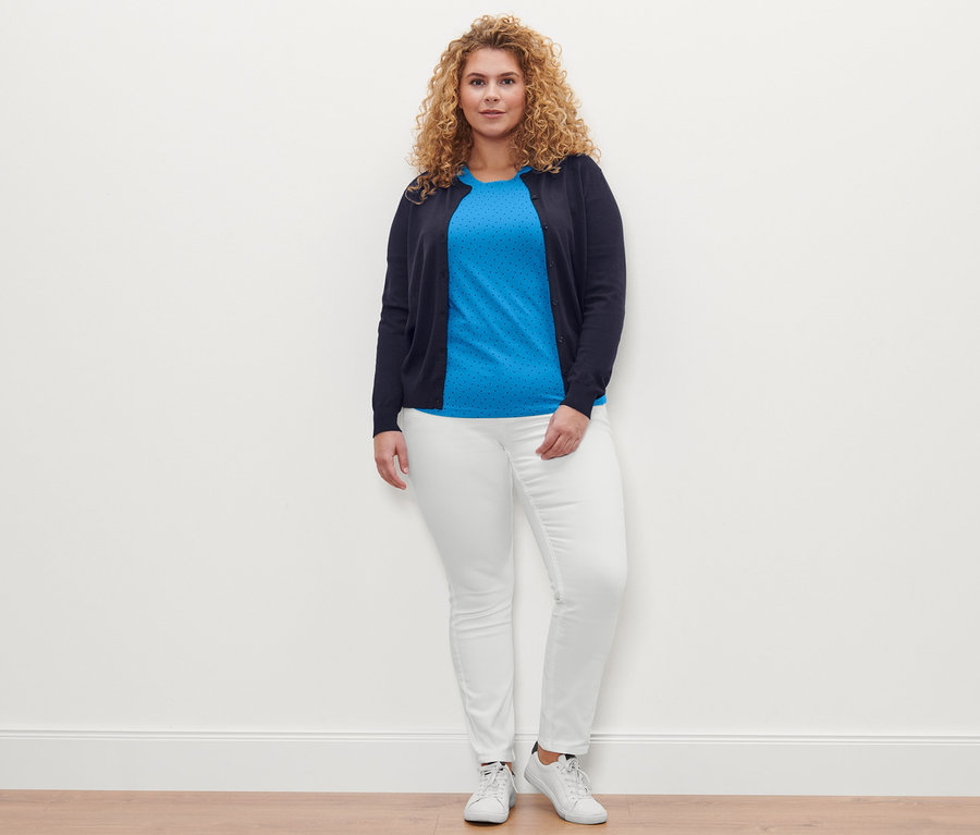 Žena pózuje na sobě má tričko s puntíky, barevné džíny »Fit Emma« smetanově bílé a kardigan z jemného úpletu.