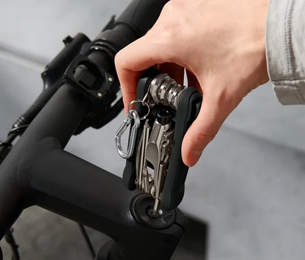 Ruka drží cyklistické multifunkční nářadí na řídítkách kola. V pozadí je vidět helma.