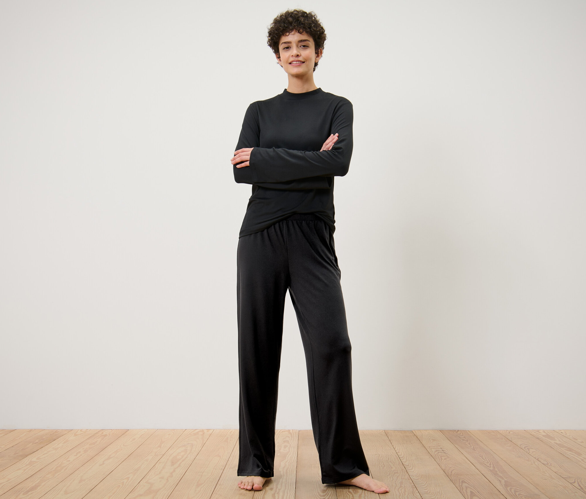 Žena stojí s rukama zkříženýma na prsou a má na sobě černé široké sportovní kalhoty.