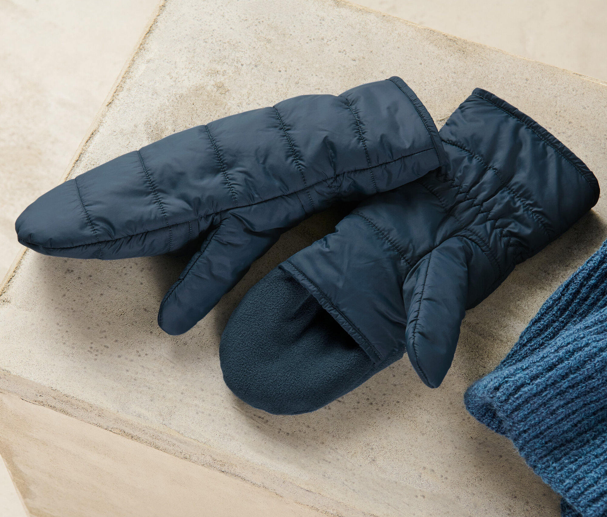 Dvoje prošívané odklápěcí palčáky tmavě modré barvy leží na šedém povrchu vedle modré pletené čepice.