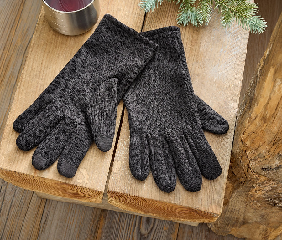 Pár šedých rukavic leží na dřevěném povrchu vedle stříbrného hrnku a větvičky jehličnanu.
