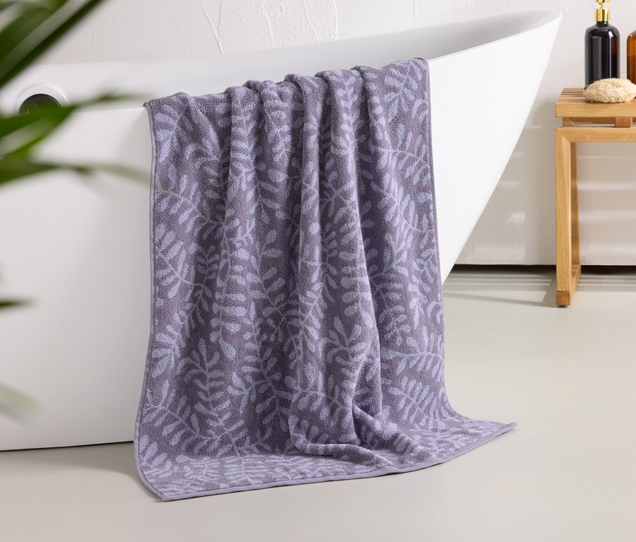 Na okraji bílé vany visí fialový ručník s potiskem listů.