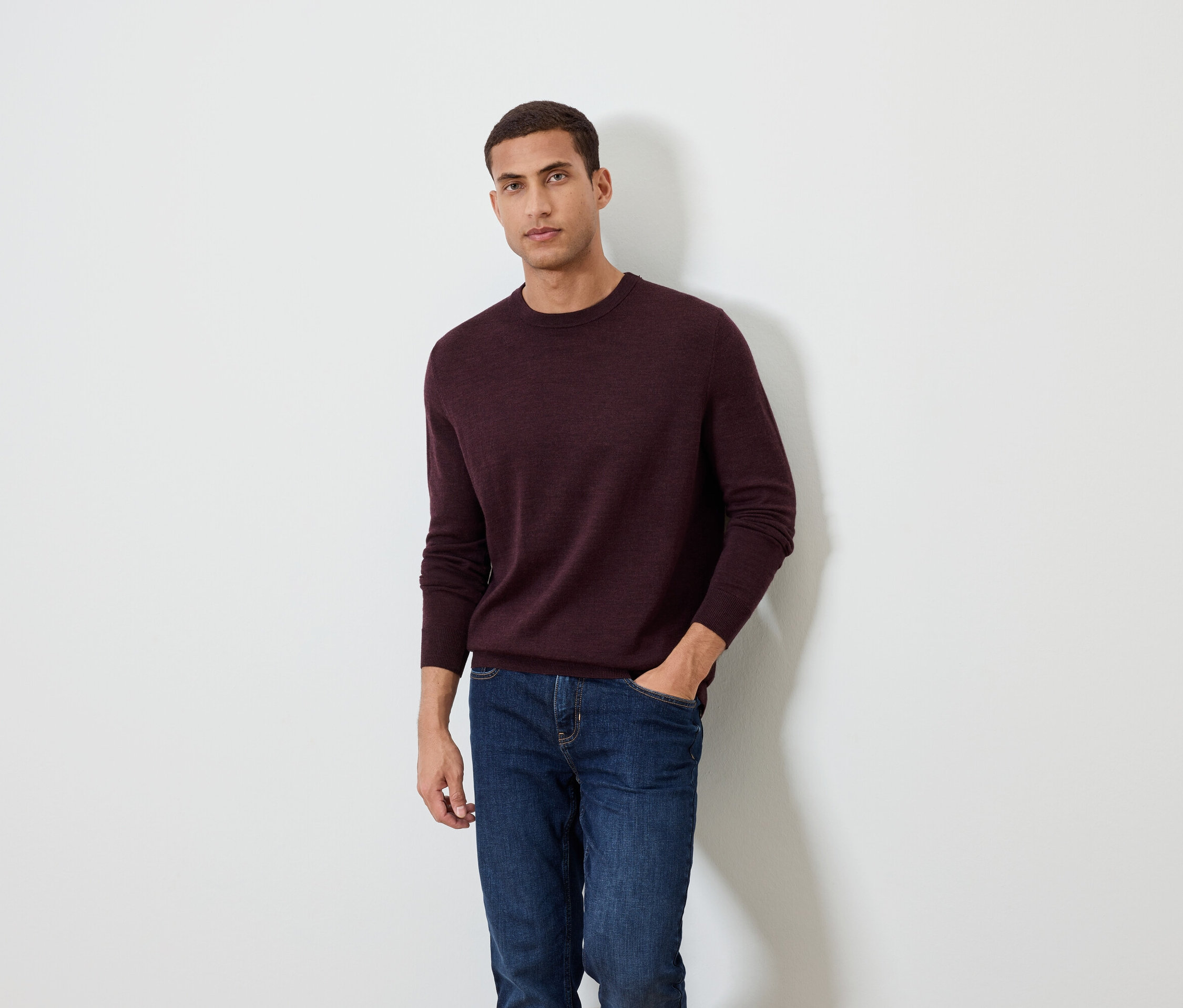 Muž pózuje na bílém pozadí a má na sobě vínový svetr a džíny.