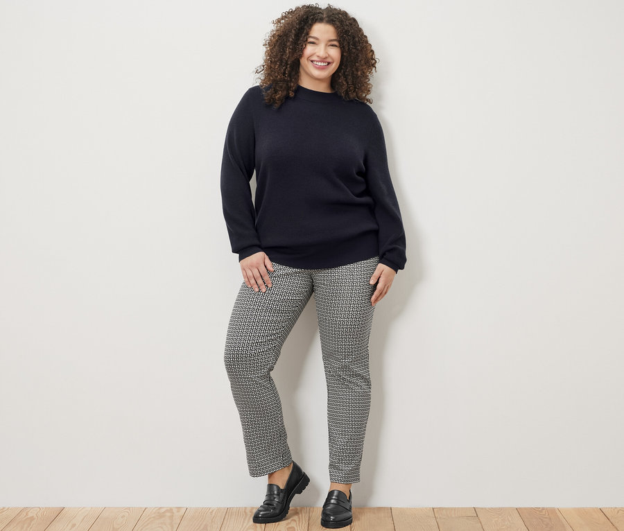 Usmívající se žena pózuje v tmavě modrém svetru a vzorovaných strečových kalhotách.