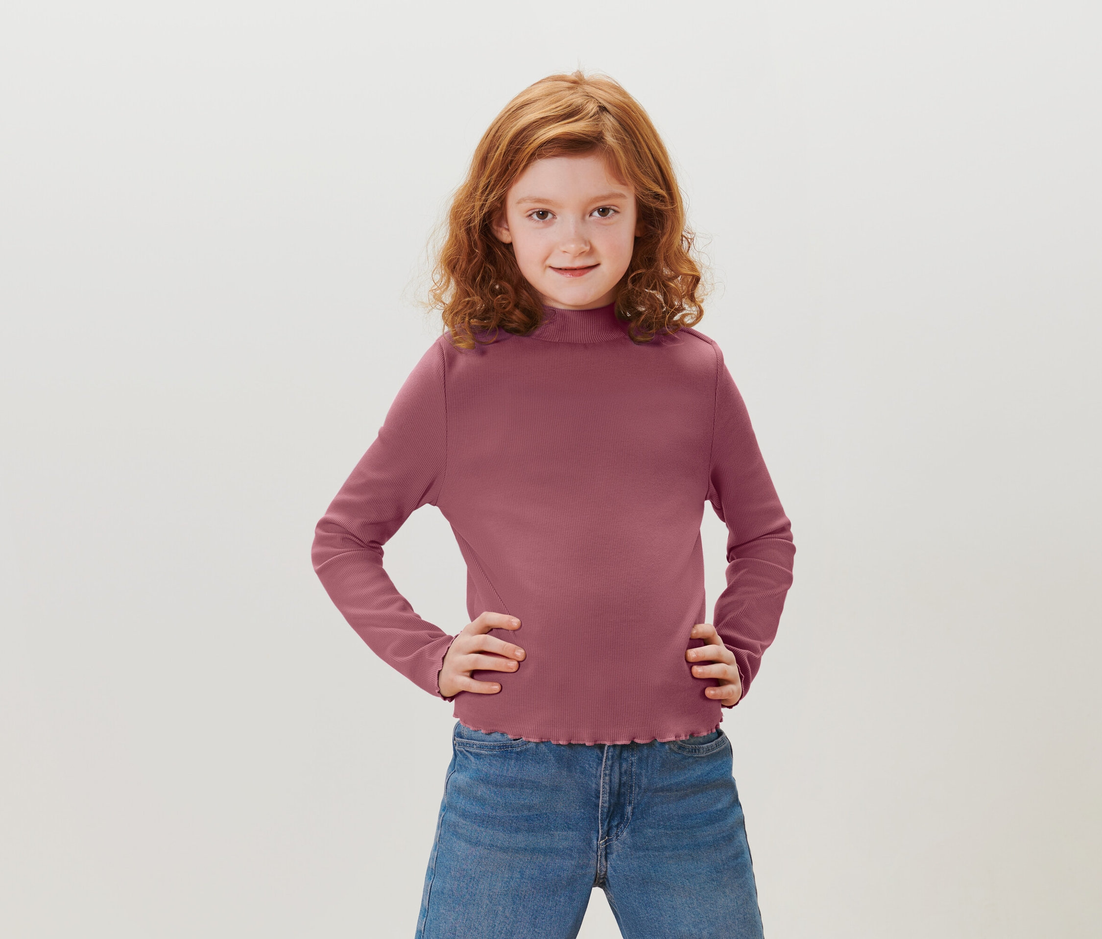 Dívka s rusovlasými kudrnatými vlasy stojí s rukama v bok. Má na sobě purpurové tričko s dlouhým rukávem a modré džíny.