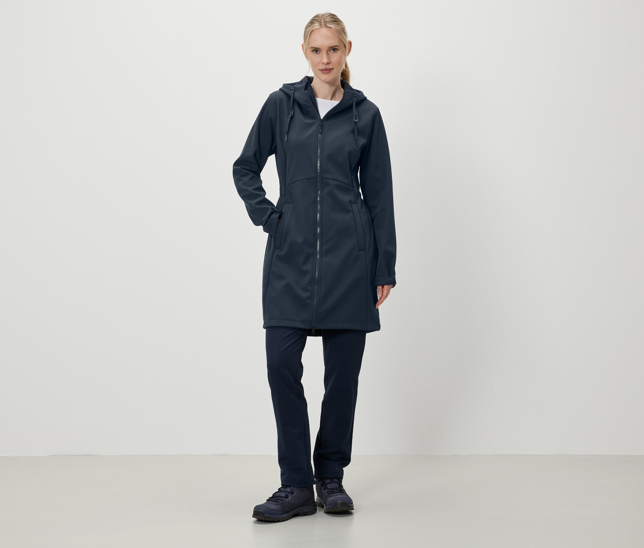 Žena stojí a má na sobě tmavě modrý kabát s kapucí, kalhoty a boty.