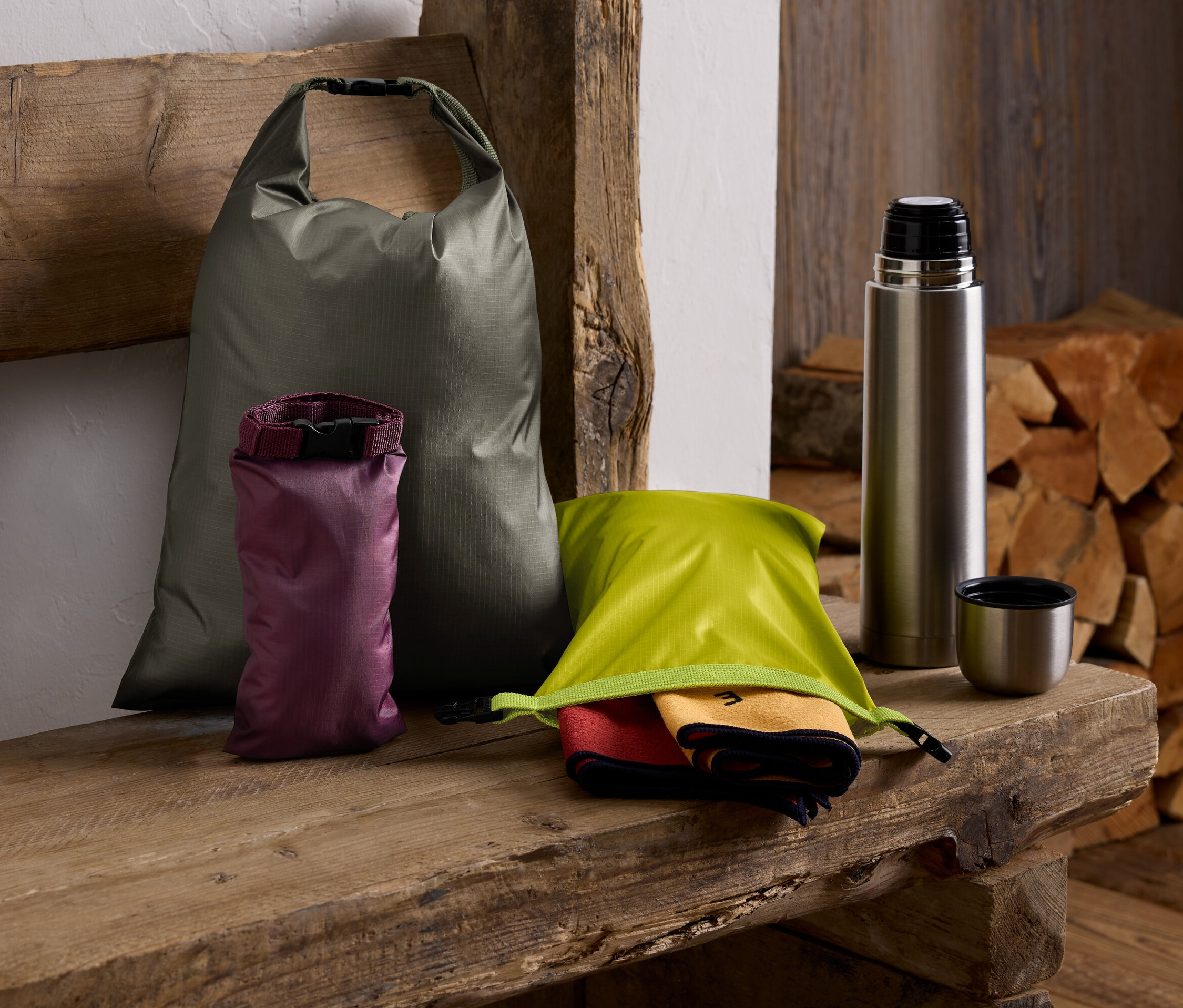 Na dřevěné lavici leží zelený vak, fialový vak, zelený vak s ručníky, termoska a štos dřeva.