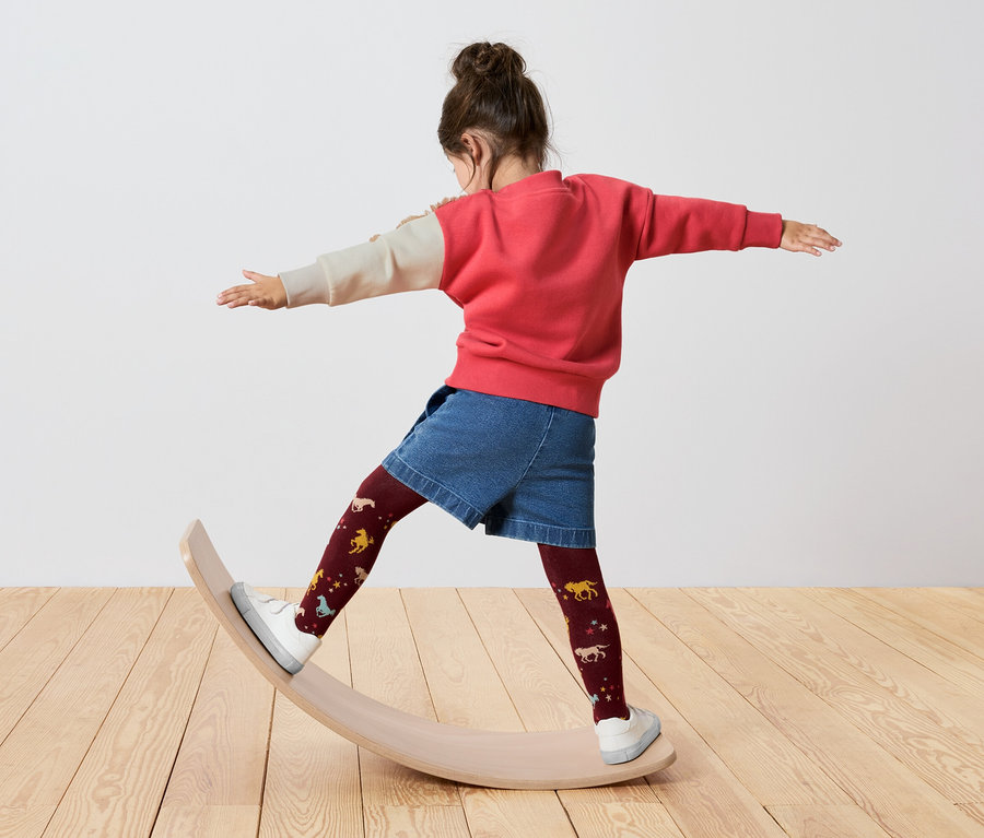 Dívka stojí na balanční desce, má na sobě červeno-krémovou mikinu, džínové kraťasy a bordó punčochy s koňmi.