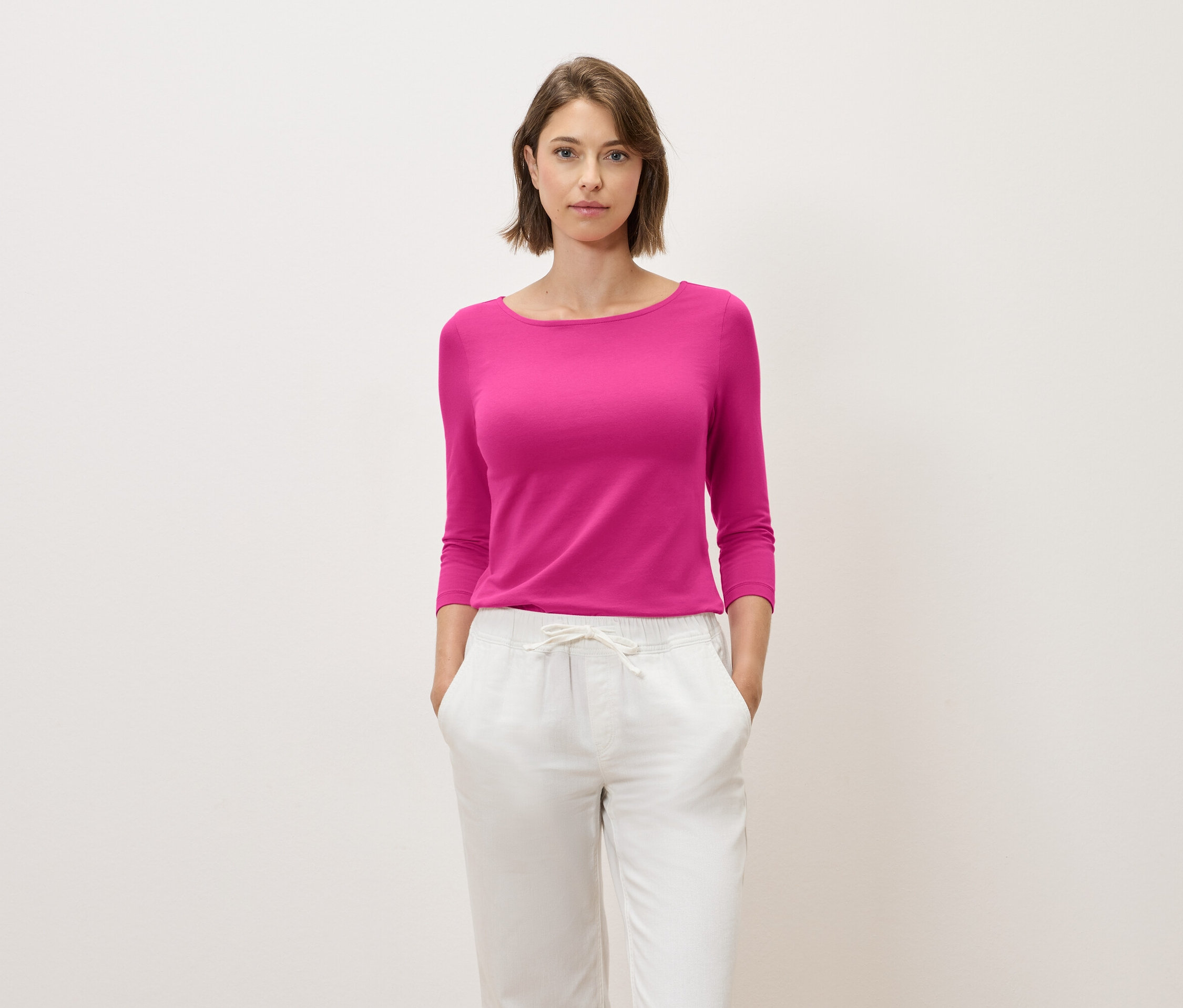 Žena s hnědými vlasy pózuje v růžovém topu s tříčtvrtečním rukávem a bílých kalhotách.