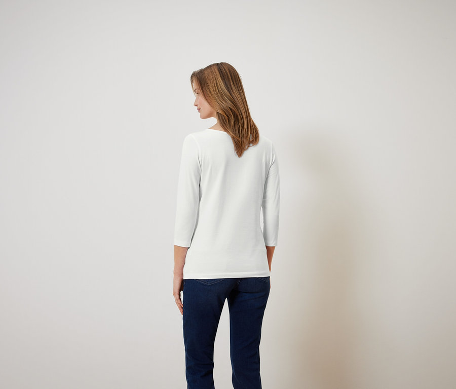 Žena s hnědými vlasy stojí zády a má na sobě bílou košili s tříčtvrtečním rukávem a modré džíny.