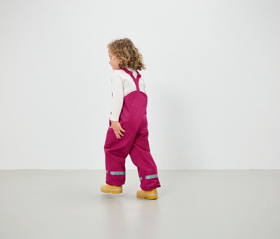 Dívka v růžových kalhotách s laclem a žlutých botách jde po podlaze.