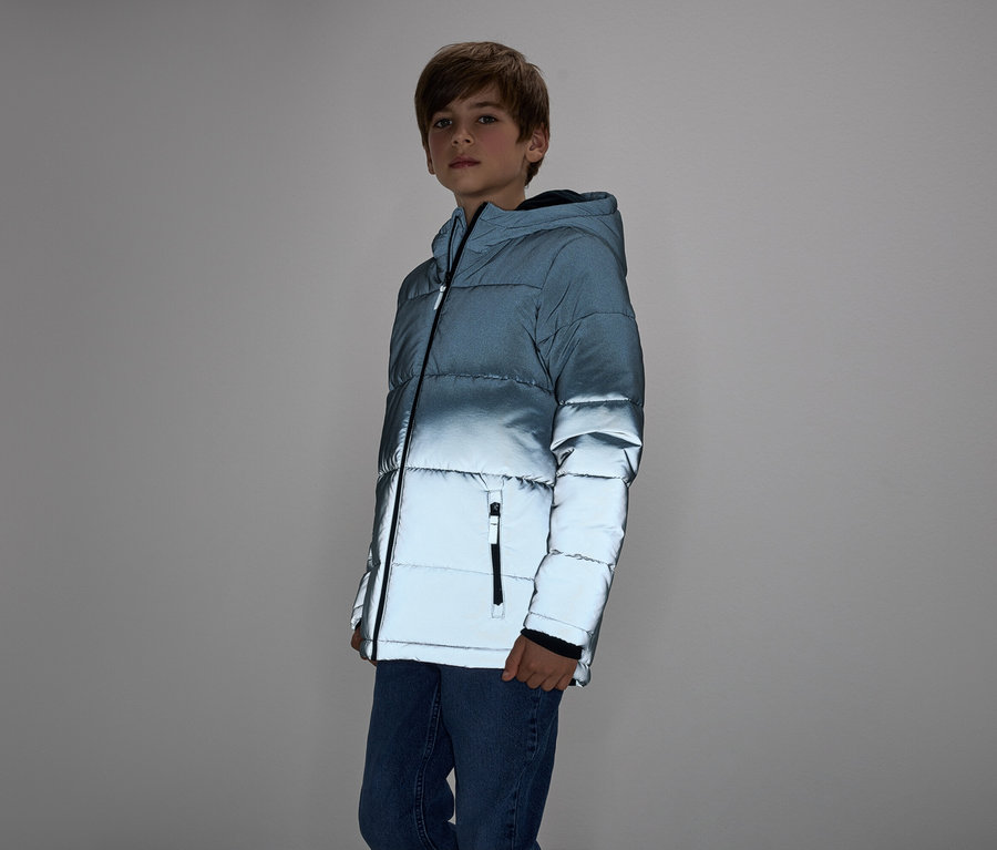 Chlapec v modré prošívané bundě a džínách stojí na šedém pozadí.