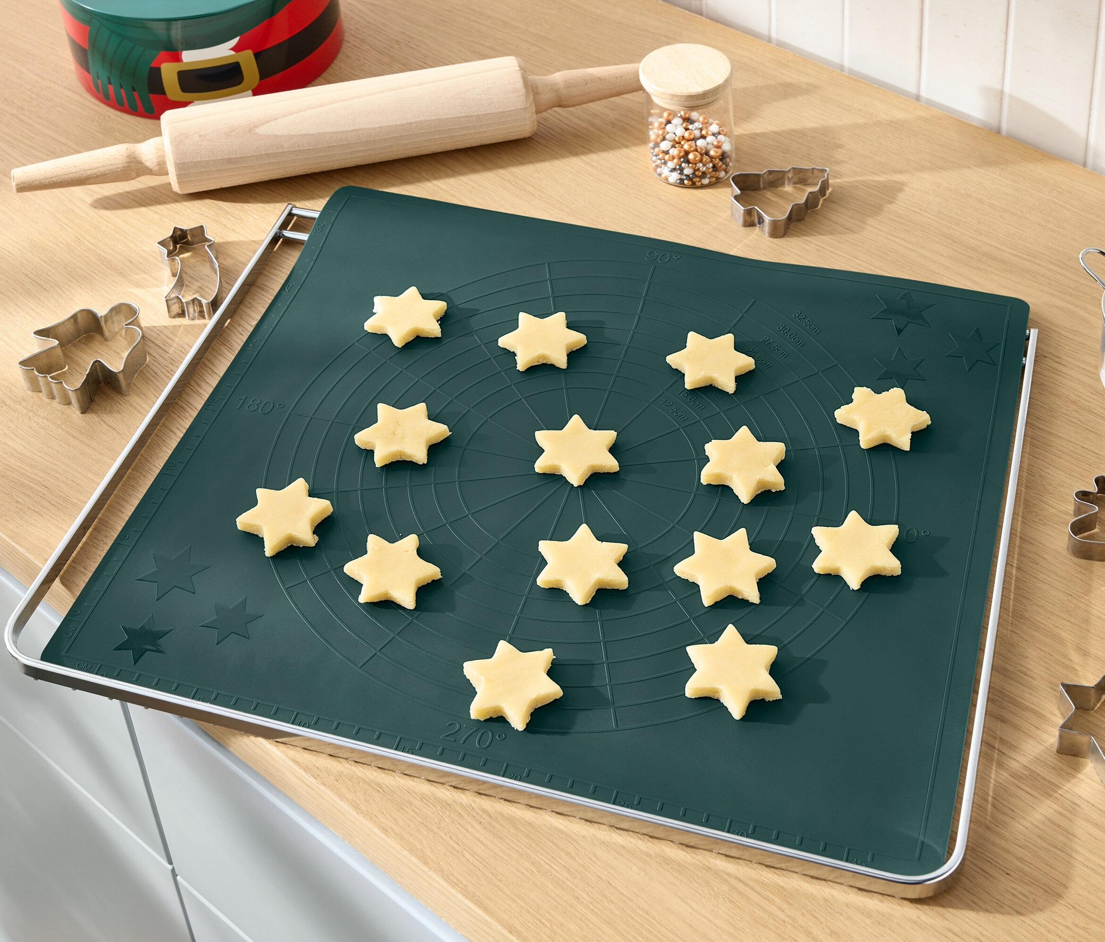 Hvězdičky z těsta na zelené podložce na pečení s válečkem, posypem a vykrajovátky na sušenky.