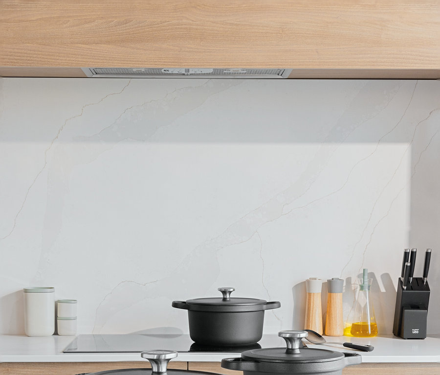 Tři černé hrnce s poklicemi na bílé kuchyňské lince. Na prkénku leží rajčata a brambory.