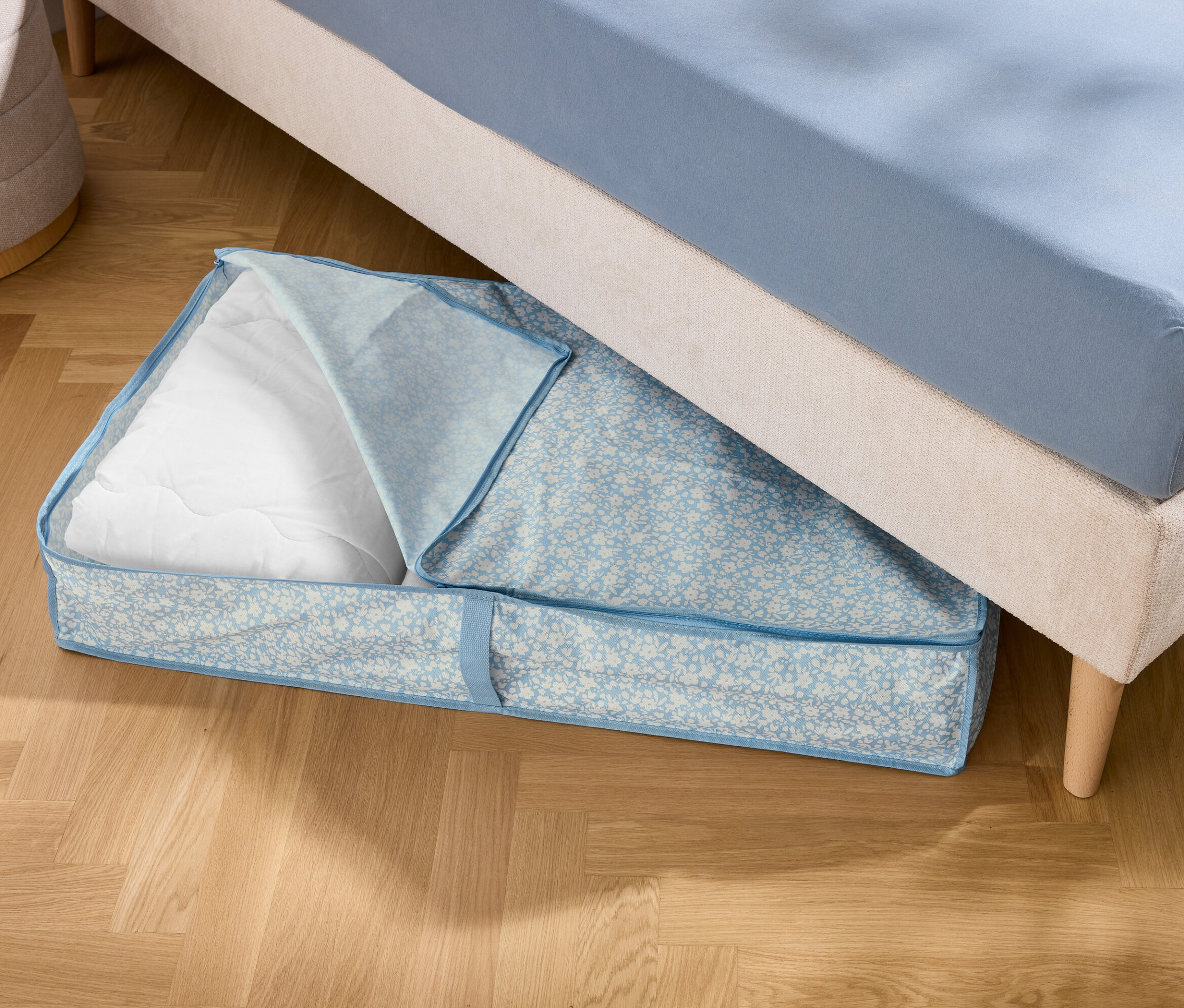 Pod postelí leží modrá úložná taška s bílou výplní.