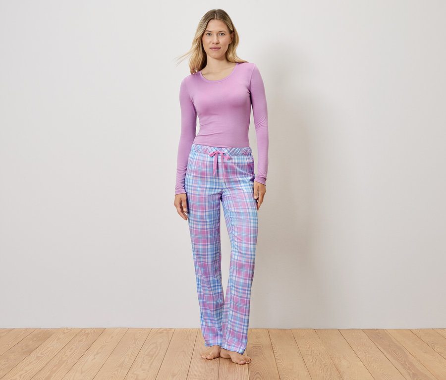 Žena v lila topu a kostkovaných pyžamových kalhotách stojí na dřevěné podlaze.
