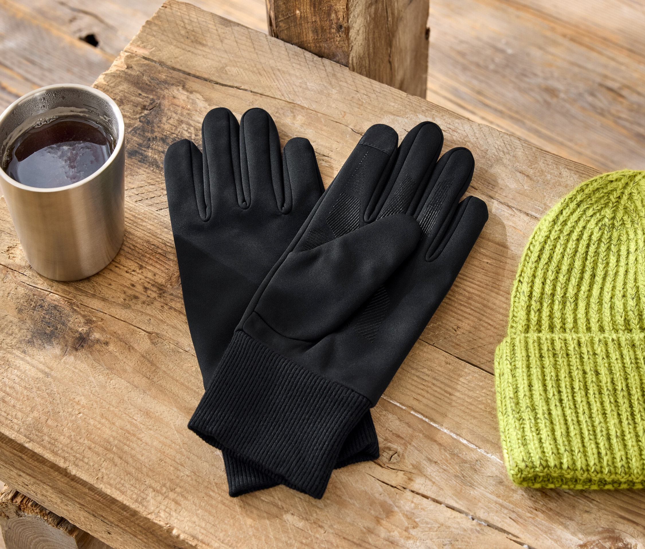 Na dřevěném stole stojí kovový hrnek, černé rukavice a zelená pletená čepice.