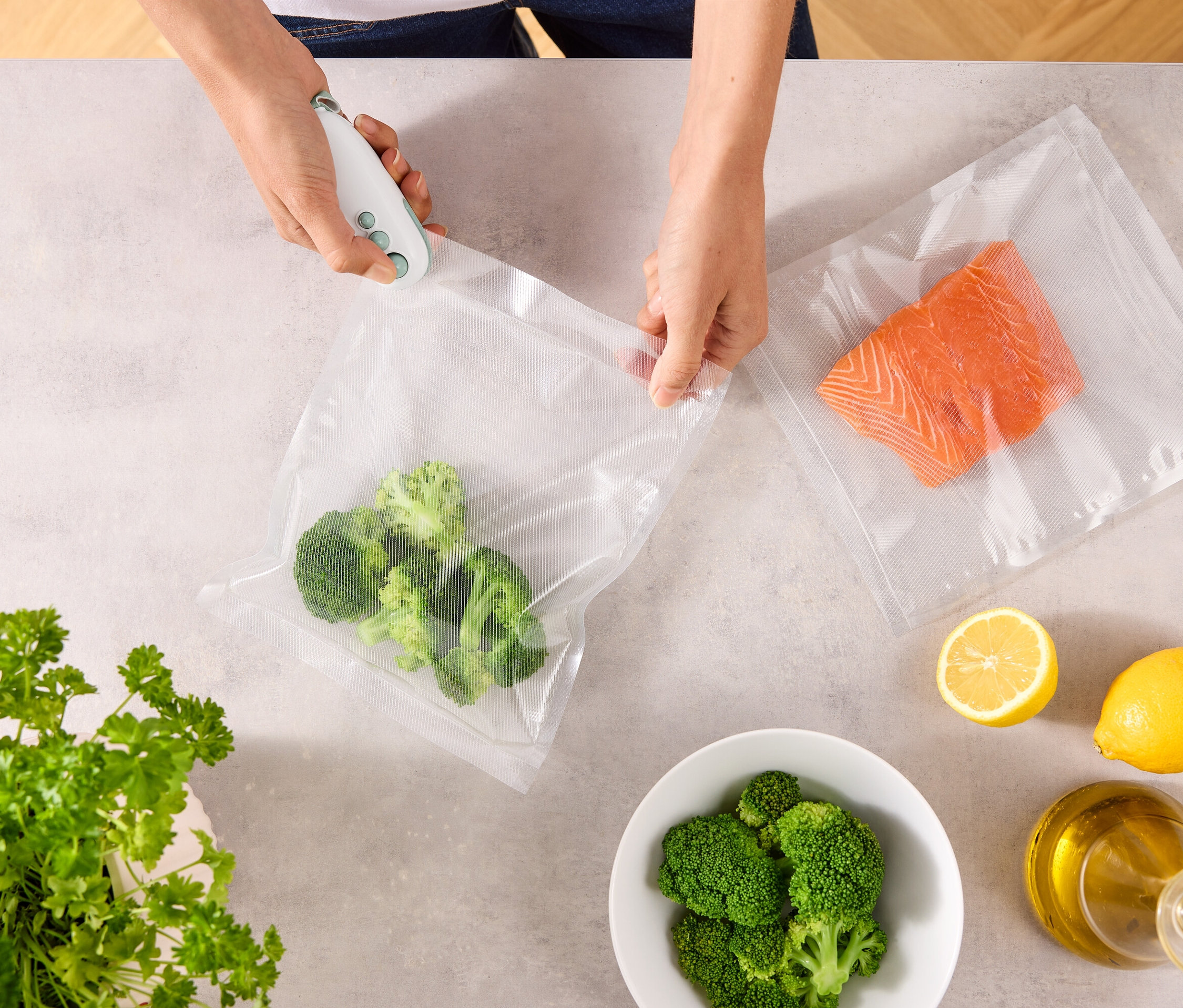 Osoba uzavírá sáček s brokolicí pomocí vakuovačky. Na stole leží další sáček s lososem, citrony, miska brokolice, petržel a láhev oleje.