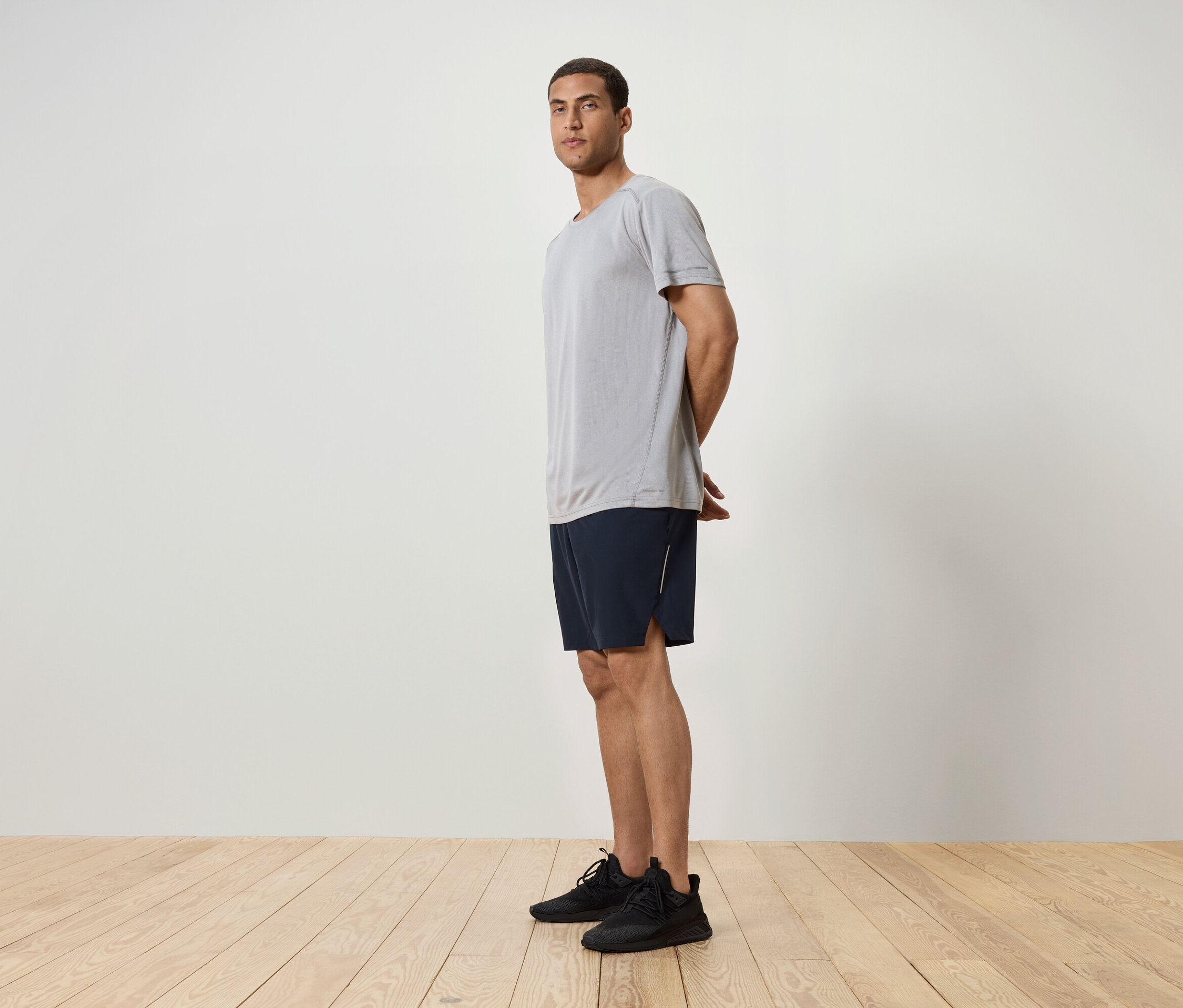 Muž stojí bokem, má na sobě šedé tričko, tmavě modré šortky a černé tenisky.