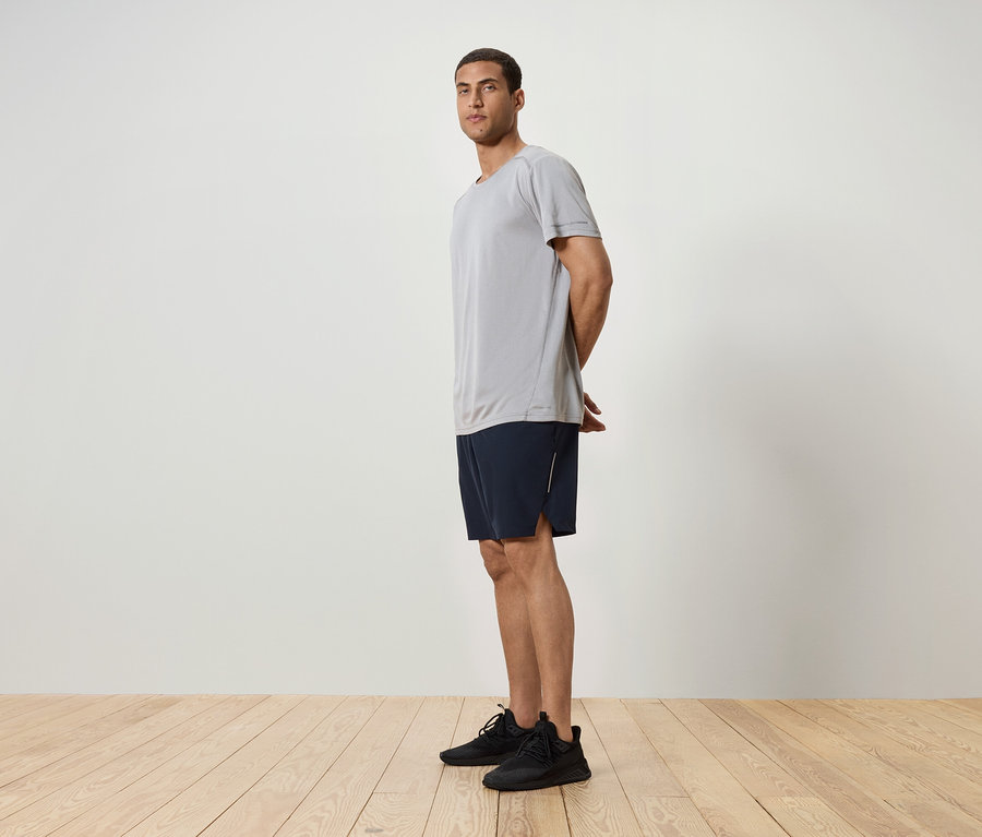 Muž stojí bokem, má na sobě šedé tričko, tmavě modré šortky a černé tenisky.