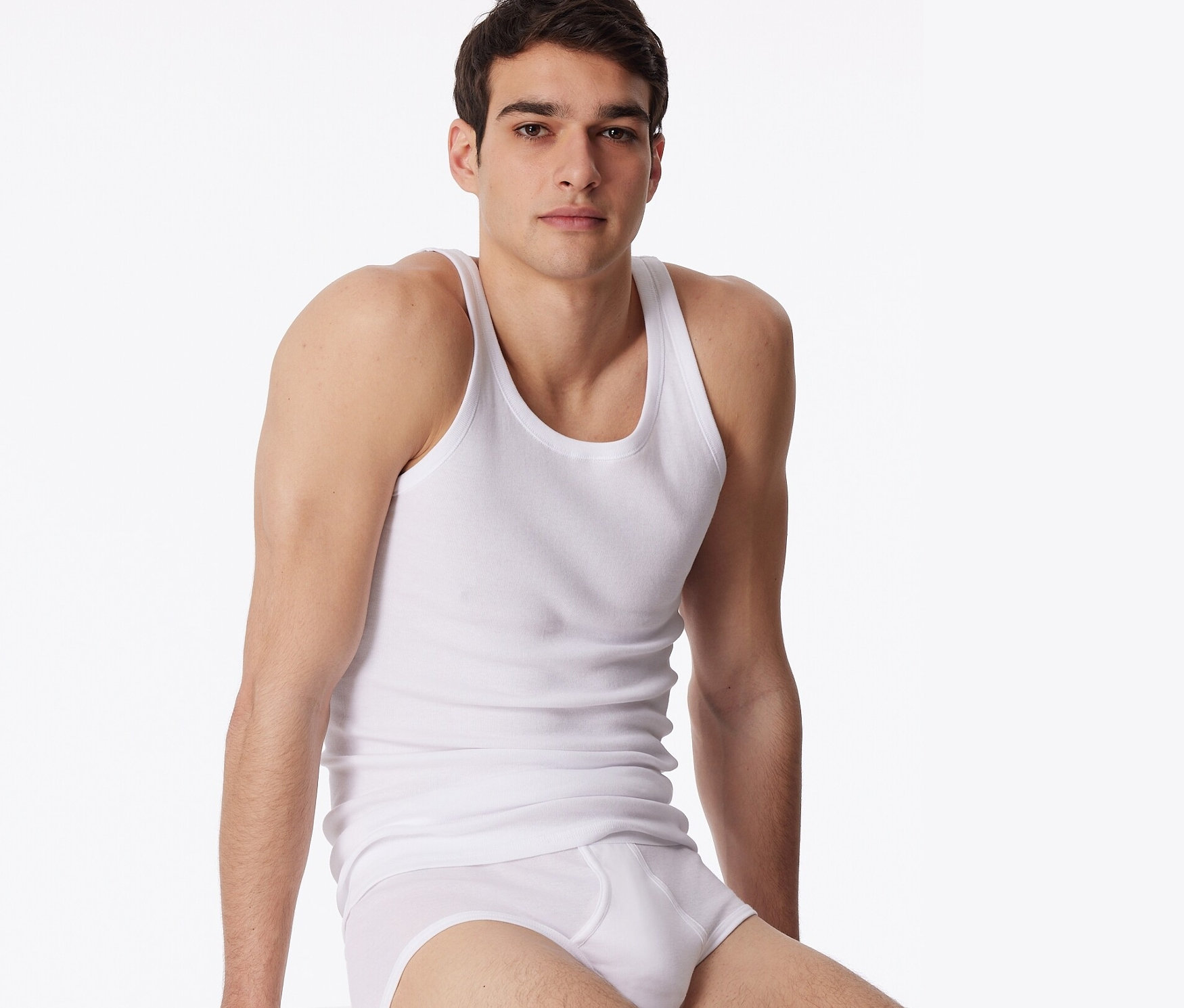 Muž sedí na bílé krychli a má na sobě bílé spodní prádlo a bílé tílko SCHIESSER z jemného žebrovaného úpletu, 2 ks.