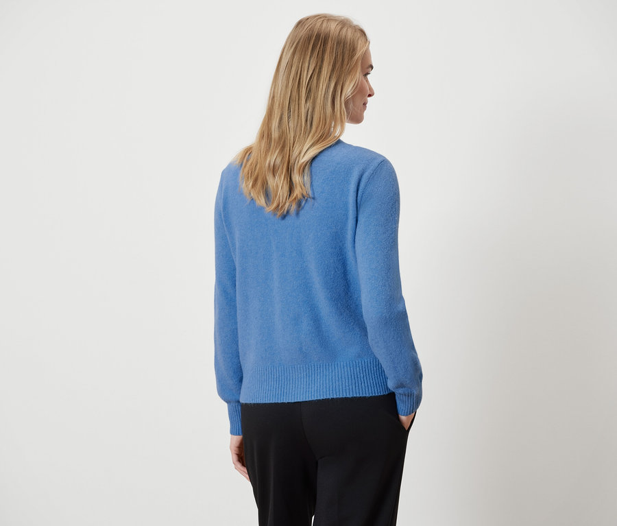 Zobrazena žena zezadu, má na sobě modrý svetr a černé kalhoty.