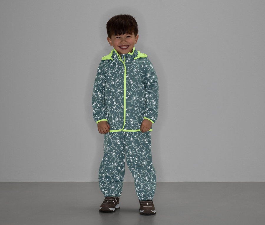Chlapec v šedé bundě a kalhotách se vzorem medvěda a neonově žlutým lemováním se usmívá.
