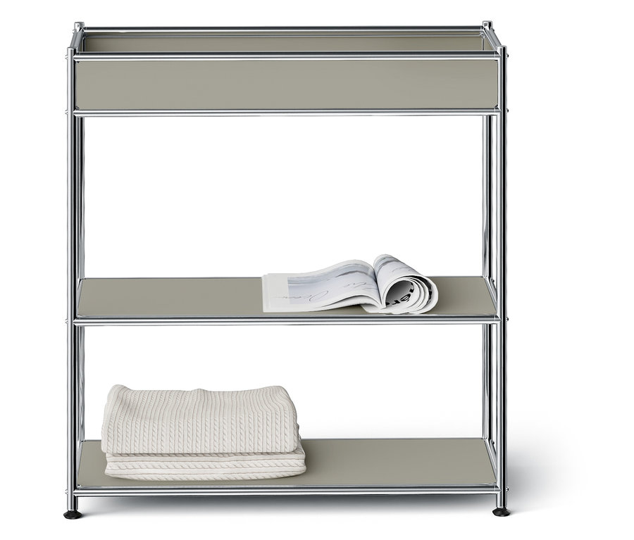 Třípatrový regál se stříbrným rámem a šedými policemi. Na prostřední polici leží otevřený časopis, na spodní polici je složená bílá deka.