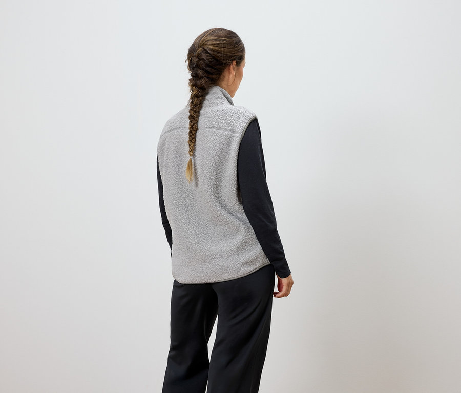 Žena zády k fotoaparátu má na sobě šedou vestu a černé kalhoty.