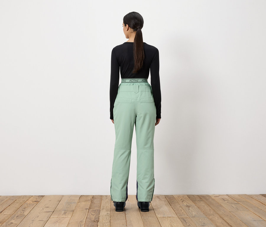 Žena zády k fotoaparátu má na sobě černé tričko s dlouhým rukávem a světle zelené kalhoty.