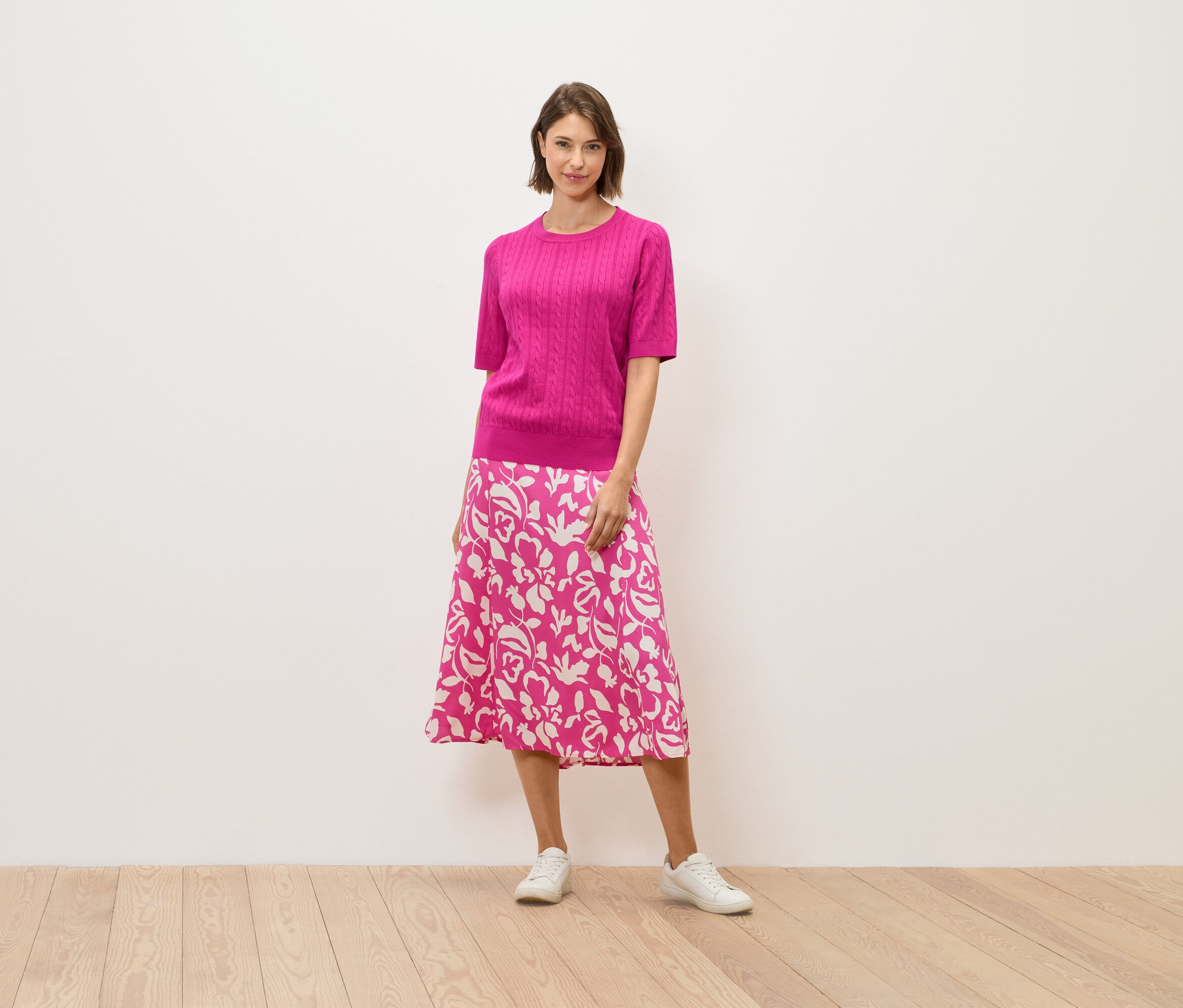 Žena v růžovém svetru a sukni s květinovým vzorem stojí na dřevěné podlaze.