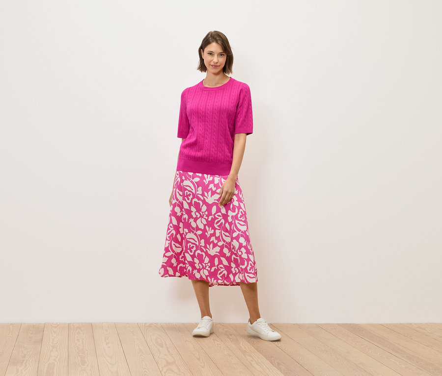 Žena v růžovém svetru a sukni s květinovým vzorem stojí na dřevěné podlaze.