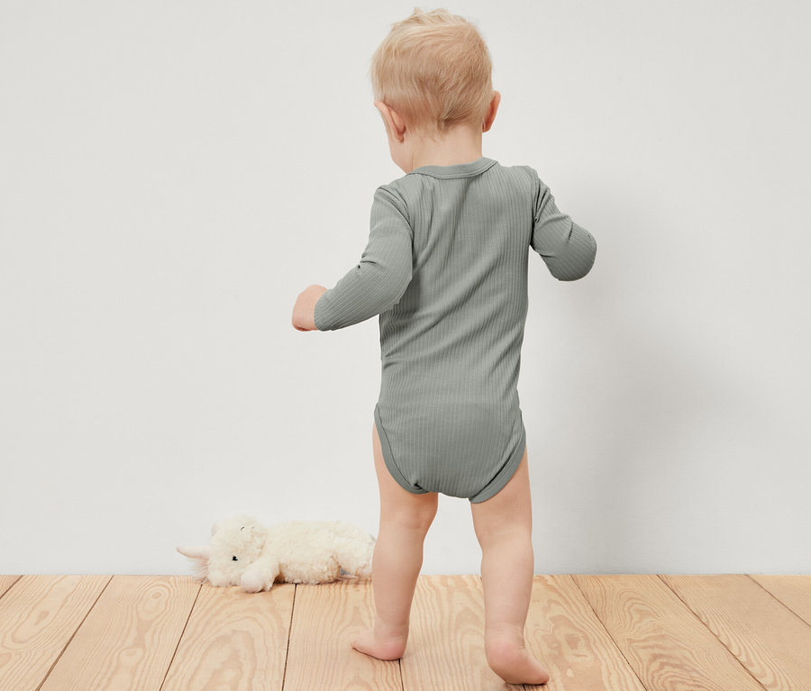 Malé dítě v žebrovaném body s dlouhým rukávem stojí na dřevěné podlaze vedle plyšového zvířete.