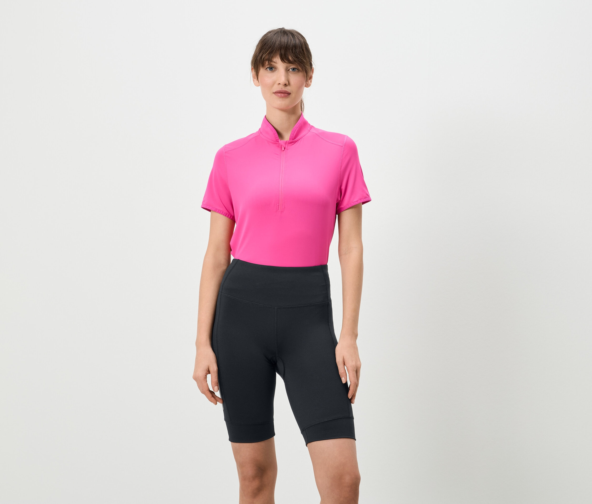 Žena stojí na bílém pozadí a má na sobě růžové tričko s krátkým zipem a krátké tmavé legíny.