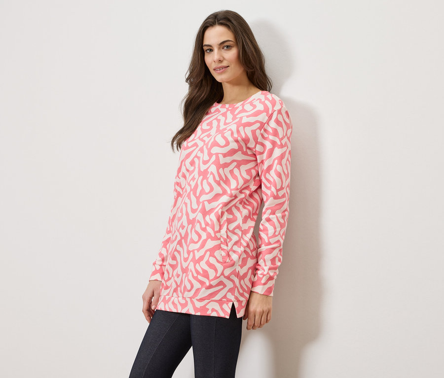 Žena má na sobě růžový svetr s abstraktním vzorem a tmavé džíny na bílém pozadí.