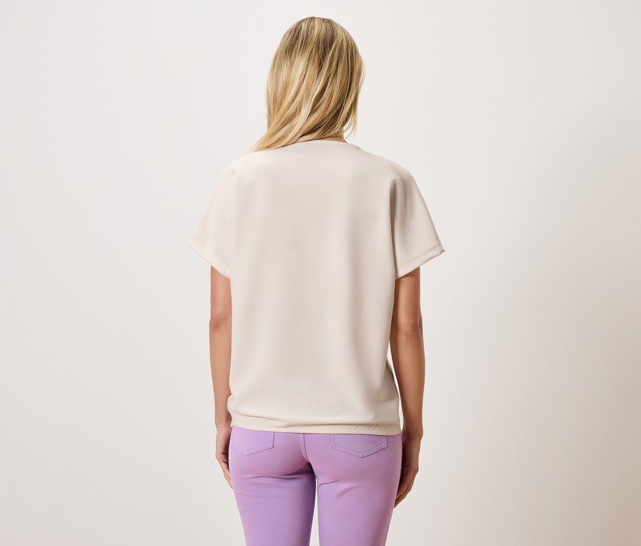 Pohled zezadu na ženu s blond vlasy, oblečenou v béžové halence a fialových kalhotách.