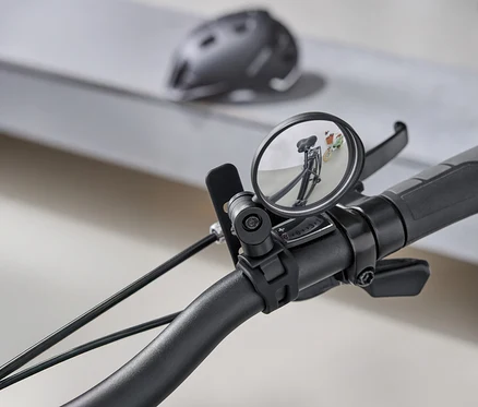 Detailní záběr na řídítka kola s kulatým zpětným zrcátkem, které odráží silnici za kolem. V pozadí je šedá cyklistická helma.