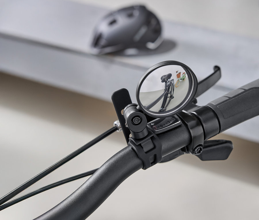 Detailní záběr na řídítka kola s kulatým zpětným zrcátkem, které odráží silnici za kolem. V pozadí je šedá cyklistická helma.